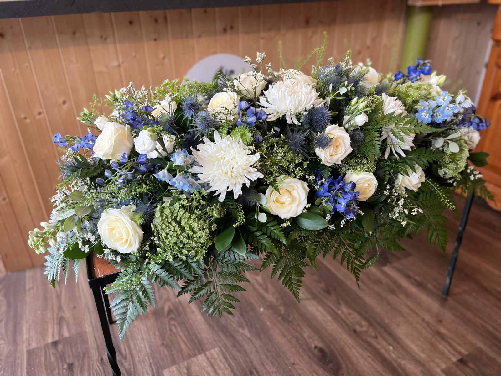 A blue and white casket spray with cream roses and seasonal greenery